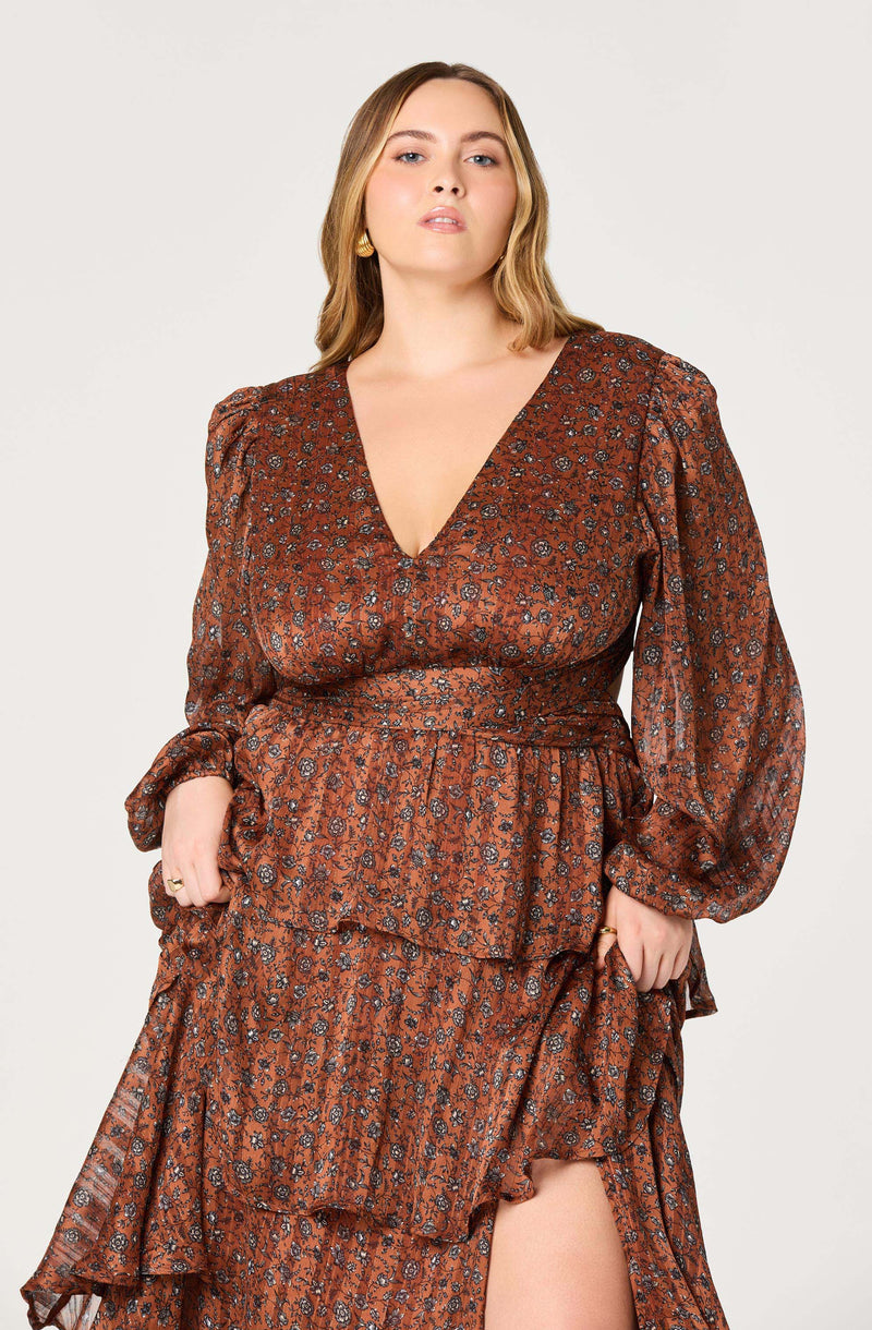 A woman is wearing a long, brown dress with a floral pattern. The dress features a deep V neckline, puffed long sleeves, and a layered, ruffled skirt with a slit revealing her leg. The fabric has a shiny, satin-like appearance.