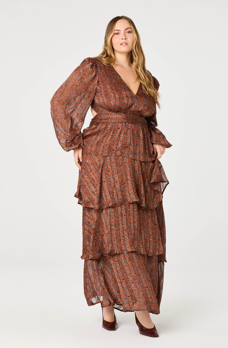 A woman is wearing a long, flowy maxi dress with a brown base color and a small, intricate floral pattern. The dress features a deep V-neckline, puffed long sleeves with elastic cuffs, and a fitted waist. The skirt has multiple layers of ruffles, creating a tiered effect that extends to the floor. She is also wearing pointed-toe high heels in a matching dark maroon shade.