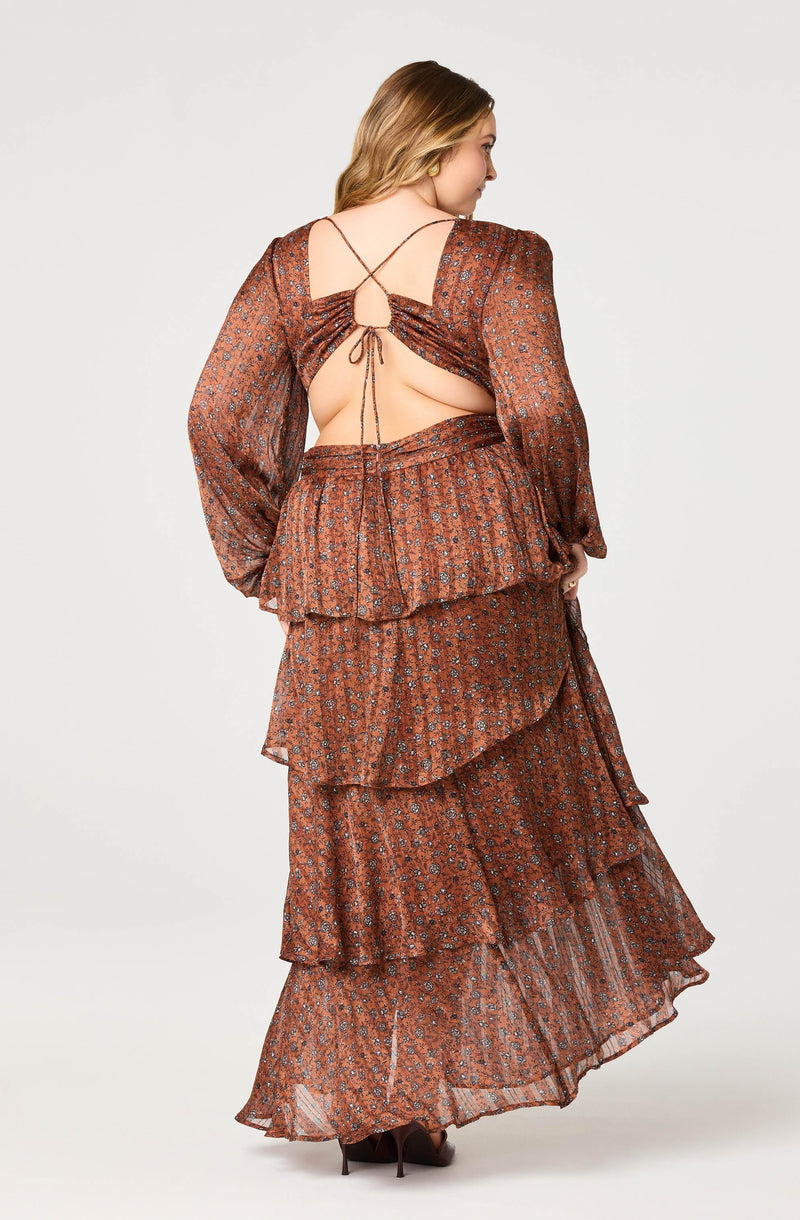A woman is wearing a long, flowy dress made of a sheer, printed fabric in shades of brown and rust. The dress features a tiered, layered design with ruffles and an open back. The top has long, slightly puffed sleeves with a subtle balloon shape. The back of the dress has a crisscross tie detail that forms a small keyhole opening. She is also wearing pointed-toe high heels.