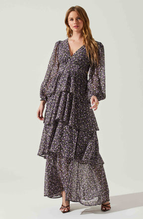 A woman is wearing a long, flowing dress made of sheer fabric with a small floral pattern in purple, white, and black. The dress features a V-neckline, long puffed sleeves with elastic cuffs, and a tiered, ruffled skirt that reaches the floor.