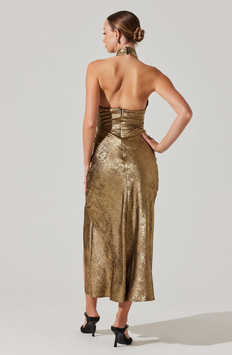 A woman is wearing a metallic gold strapless gown with a fitted bodice and a high neckline. The dress is long, reaching down to her mid-calf, and has a shiny, textured fabric. She is also wearing black high-heeled sandals. Her hair is styled in a sleek bun, and she has on a pair of earrings.