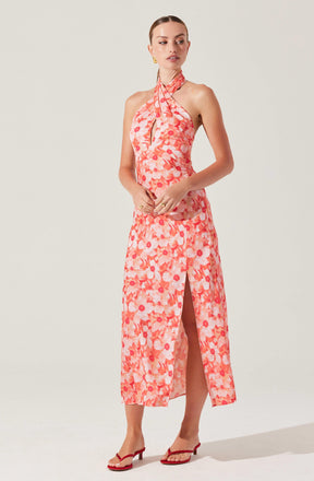 A woman is wearing a sleeveless, halter-neck maxi dress with a pink and coral floral pattern. The dress features a keyhole opening at the neckline and a front slit that reaches below the knee. She is also wearing red open-toe heeled sandals and gold earrings.