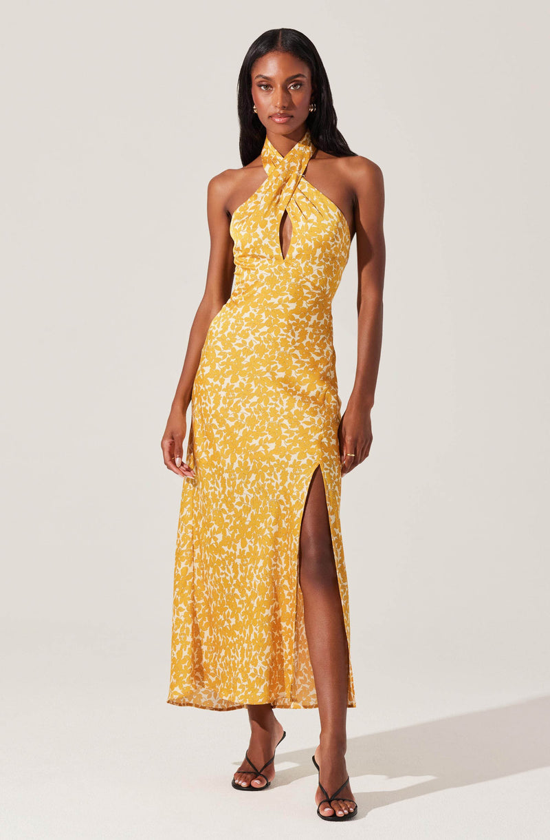 A woman is wearing a sleeveless, yellow maxi dress with a white floral pattern. The dress features a halter neckline with a keyhole cutout at the chest, and has a high slit on the side revealing her legs. She is wearing black strappy high-heeled sandals.