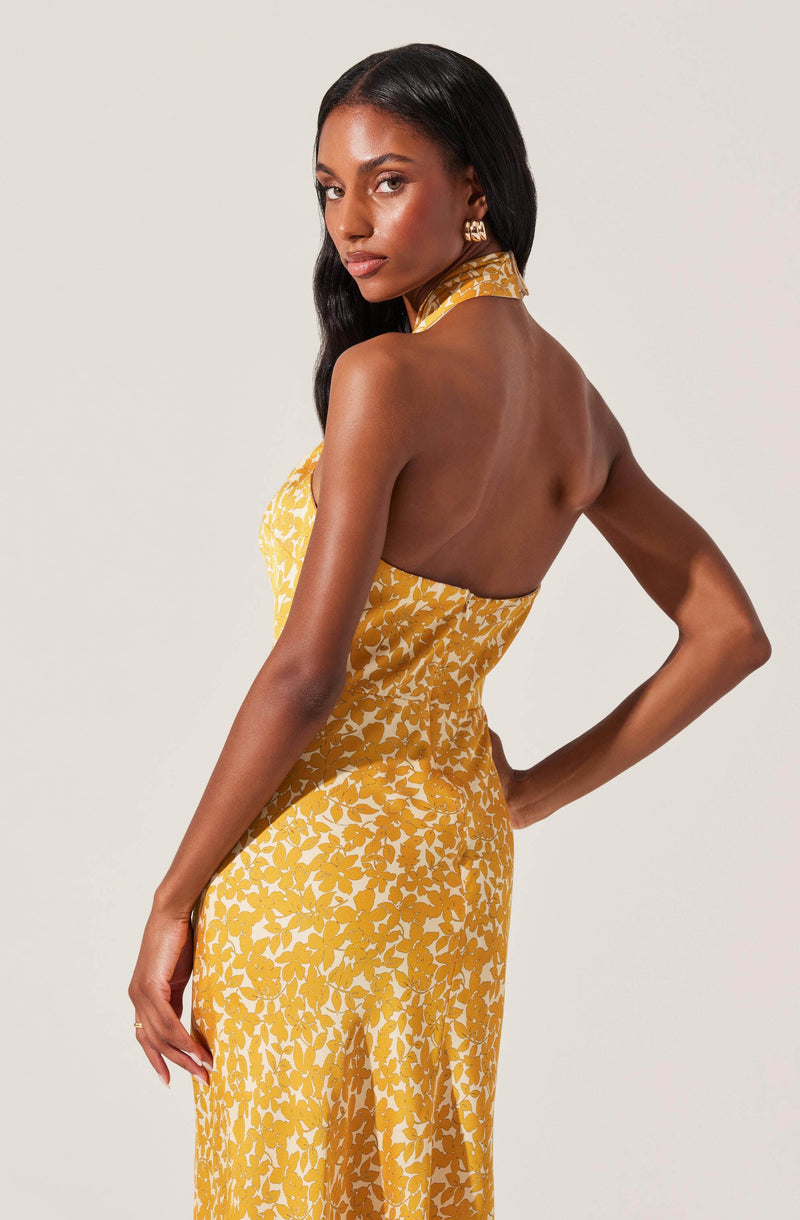 A woman is wearing a sleeveless, halter-neck dress with a yellow and white floral pattern. The dress features an open back design, and the fabric appears lightweight and flowy. She is accessorized with large gold earrings and has long, dark hair styled in loose waves.