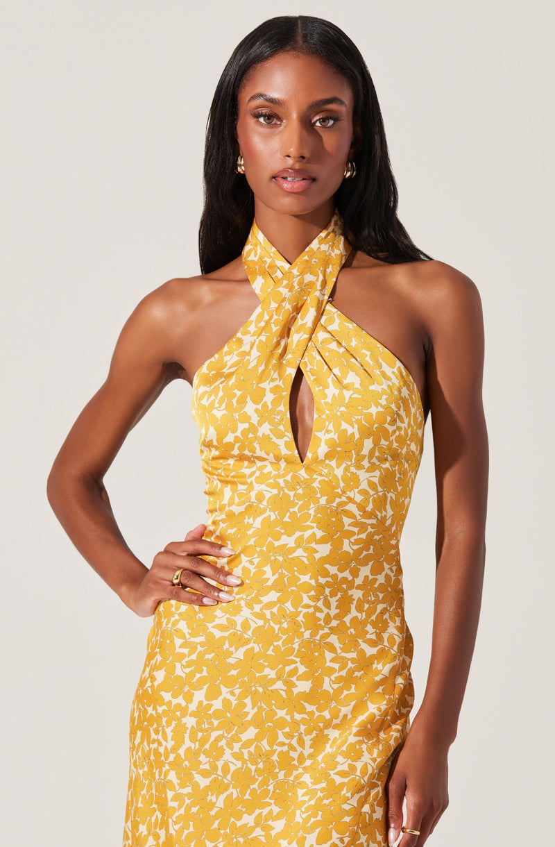 A woman is wearing a sleeveless yellow dress with a white floral pattern. The dress features a halter neck with a large tied bow at the neck, creating a keyhole opening in the front.