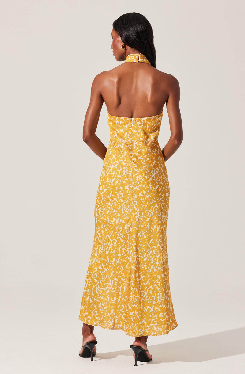 A woman is wearing a long, strapless yellow dress with a white floral pattern. The dress features a high neckline with a matching choker-style collar. She is also wearing black high-heeled sandals.