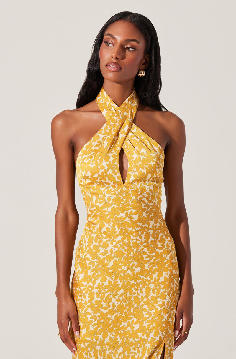 A woman is wearing a sleeveless yellow dress with a white floral pattern. The dress features a halter neck with a large bow tied at the front, creating a keyhole cutout just below the bow. The fabric is fitted and flows down in an elegant silhouette.