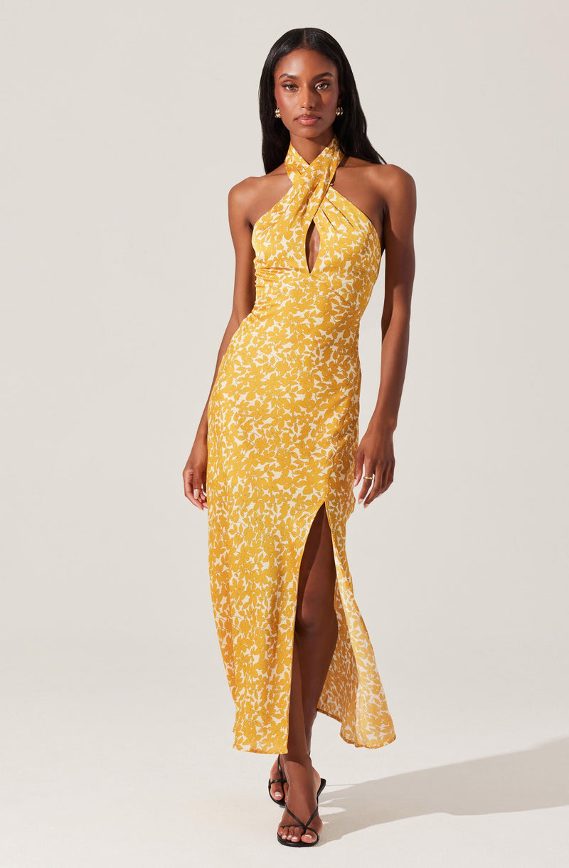 A woman is wearing a sleeveless, yellow maxi dress with a white floral pattern. The dress features a halter neck that ties into a bow at the front, with a keyhole opening below the tie, and a high slit on the left side revealing her leg. She is also wearing black high-heeled sandals.