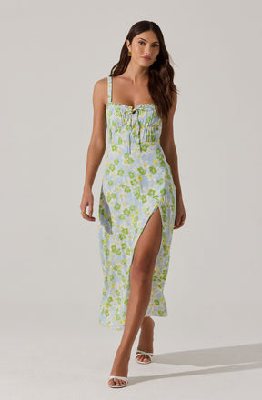 A woman is wearing a sleeveless, ankle-length dress with thin straps and a slit that reveals her leg. The dress has a light blue base color with a green and yellow floral pattern, and features a gathered, ruched bodice with a bow and tie detail at the center. She is also wearing white high-heeled sandals with an open toe.