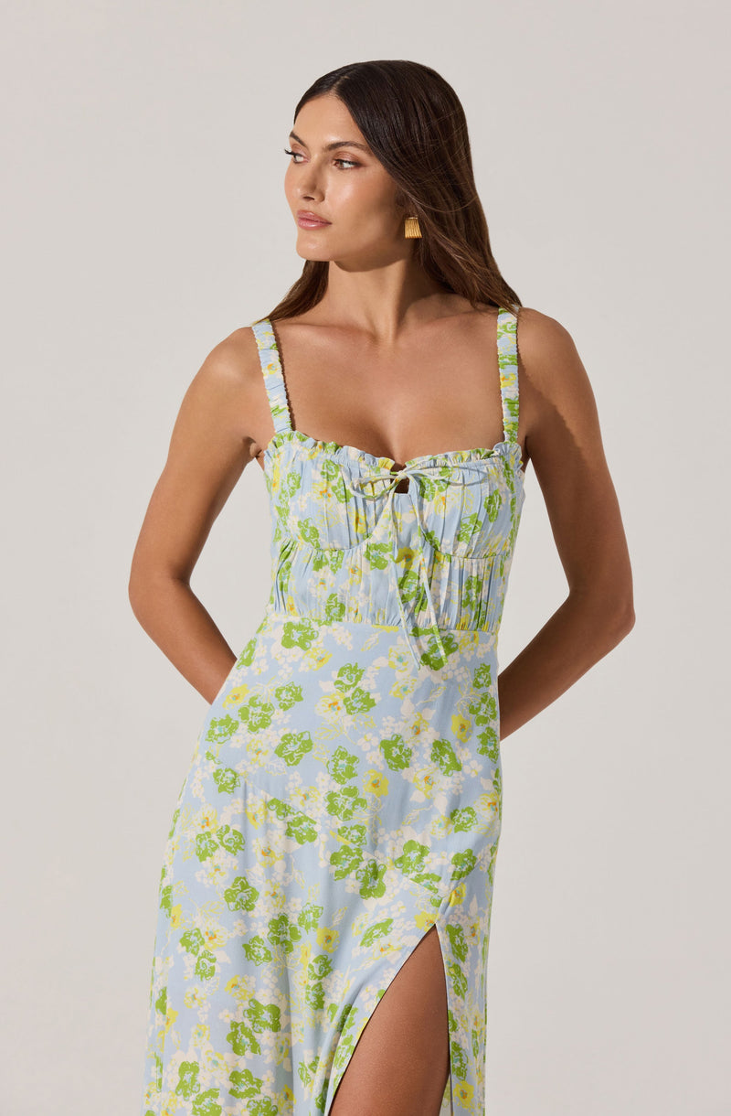 A woman is wearing a sleeveless maxi dress with a floral pattern. The dress has a light blue background with green and white flowers, features adjustable shoulder straps, a gathered bodice with a drawstring tie at the neckline, and a high slit on the side.