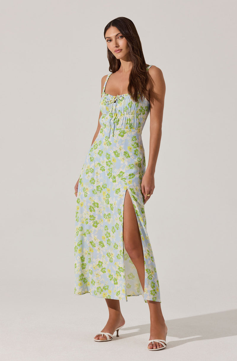 A woman is wearing a sleeveless, light-colored maxi dress with a floral pattern in shades of green and blue. The dress features thin straps, a gathered bodice with a bow at the center, and a high slit on one side. She is also wearing white heeled sandals.