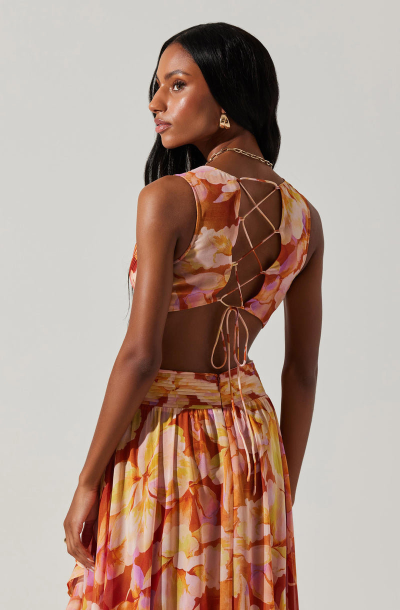 A woman is wearing a sleeveless, floral dress with shades of pink, orange, and cream. The top features a lace-up back with thin ties, creating a crisscross pattern, and the fabric appears lightweight and flowy. The skirt is gathered at the waist, with a wide waistband and a pleated, flowing design. She accessorized with a gold chain necklace and gold earrings.