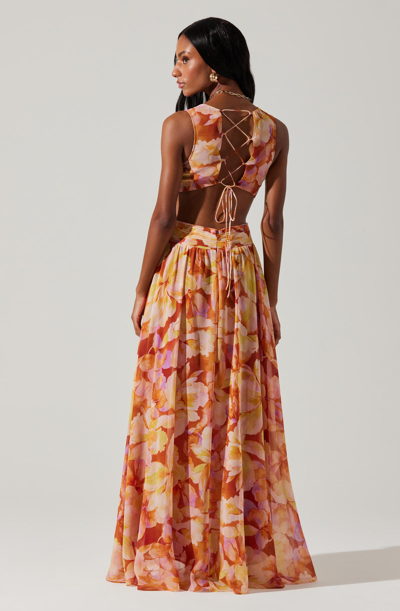 A woman is wearing a two-piece outfit consisting of a sleeveless crop top and a long, flowing skirt. The crop top features a floral pattern in shades of pink, orange, and cream, with a lace-up detail at the back that creates a crisscross pattern and ties into a bow. The skirt matches the floral pattern of the top, is high-waisted, and has a loose, billowy fit with multiple pleats that add volume and movement.