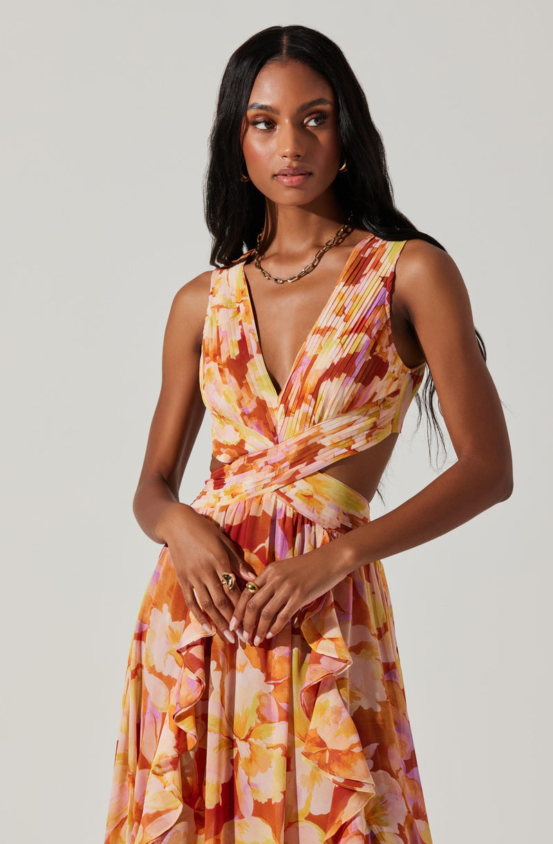 A woman is wearing a sleeveless, V-neck dress with a colorful, abstract floral pattern in shades of orange, pink, yellow, and purple. The dress features cutouts at the waist and ruffled details along the skirt.
