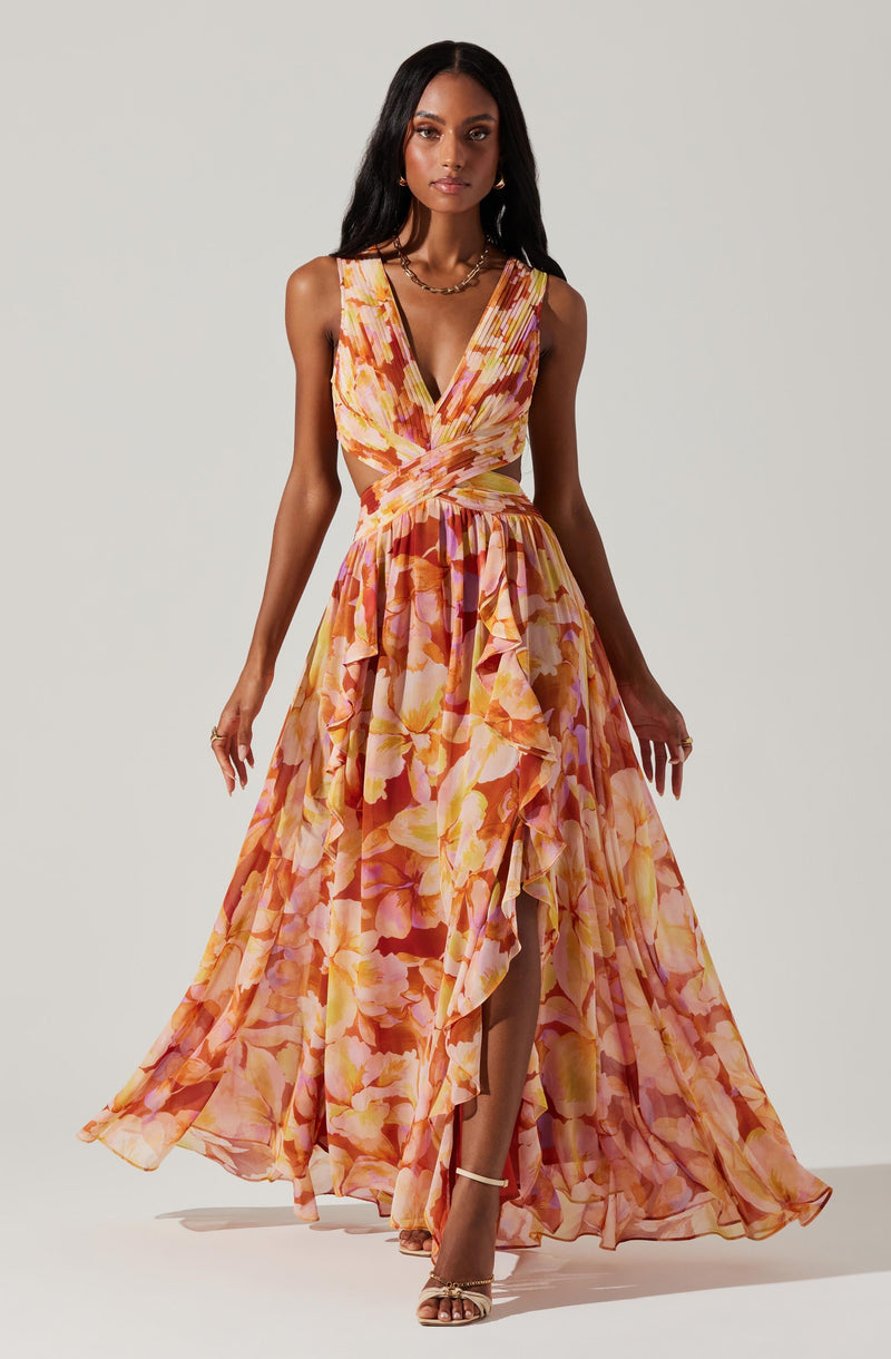 A woman is wearing a sleeveless, V-neck maxi dress with a fitted bodice and a full, flowy skirt. The dress features an abstract floral pattern in shades of pink, orange, and yellow. The skirt has a high slit at the front, revealing her legs and strappy high-heeled sandals.