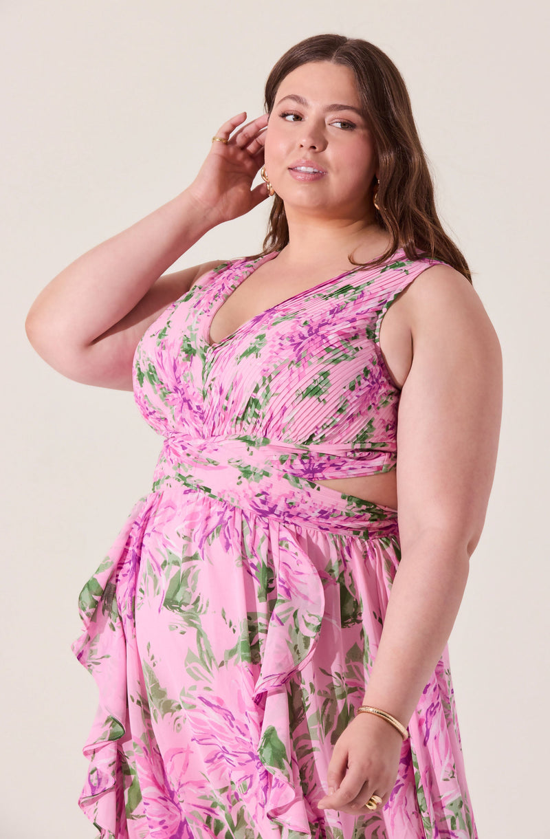 A woman wearing a sleeveless, pink floral dress with green and purple accents. The dress features a V-neckline, cut-out details at the waist, and ruffled layers on the skirt.