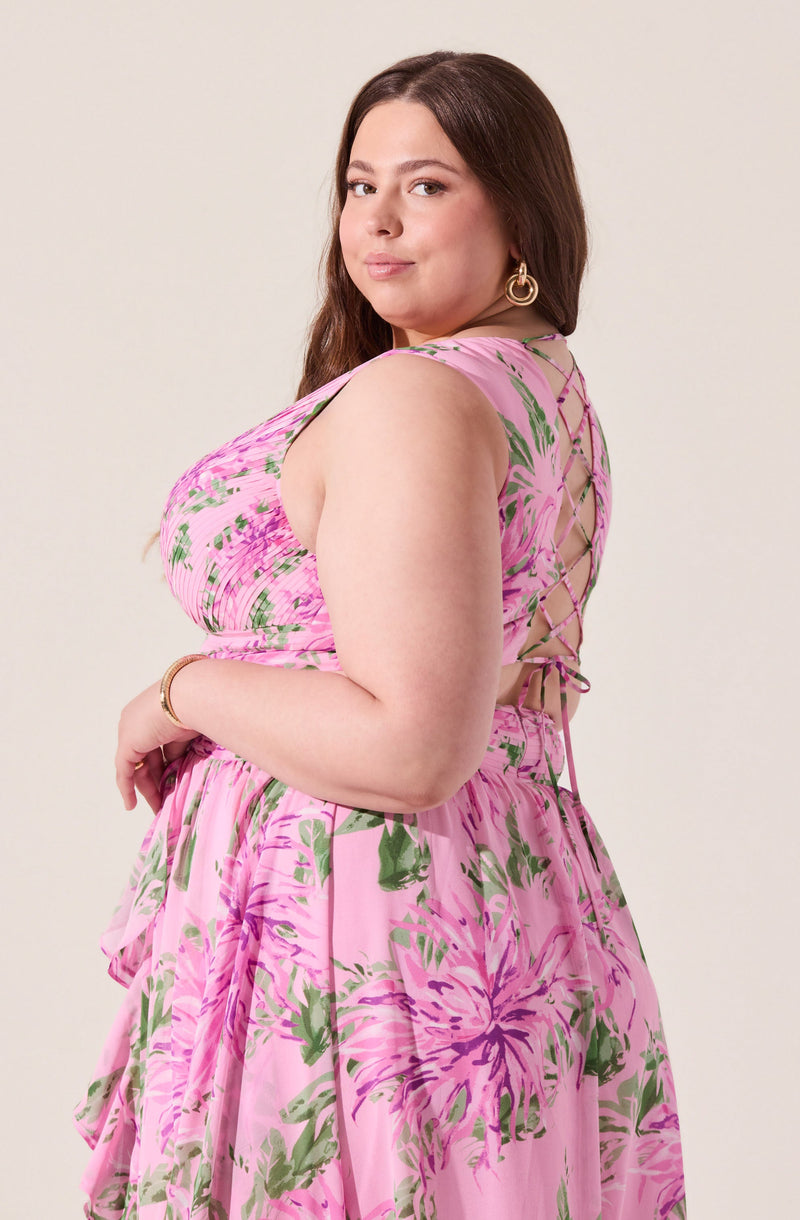 A woman is wearing a sleeveless, pink floral dress with green and purple accents. The dress features a lace-up back detail with crisscrossing ties. The fabric is lightweight and flowy, with a gathered waistline. She accessorizes with large hoop earrings and a bracelet.