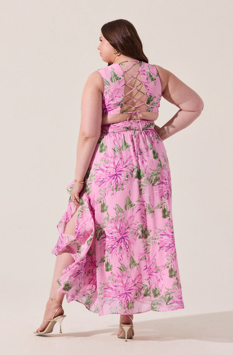 A woman is wearing a sleeveless pink dress with a floral pattern featuring large pink and green flowers. The back of the dress has a lace-up detail with crisscrossing ribbons, exposing some skin. She is also wearing a sheer, long-sleeved beige blouse underneath the dress.