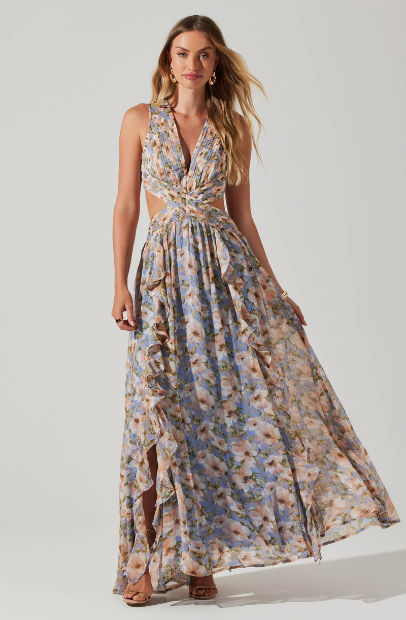 A woman is wearing a sleeveless, V-neck, floral maxi dress with a multicolored pattern. The dress features a twisted knot at the waist, ruffled details along the sides of the skirt, and a high-low hemline with a slit revealing her legs and beige heels. She has long, wavy blonde hair and is accessorized with gold hoop earrings and a bracelet.