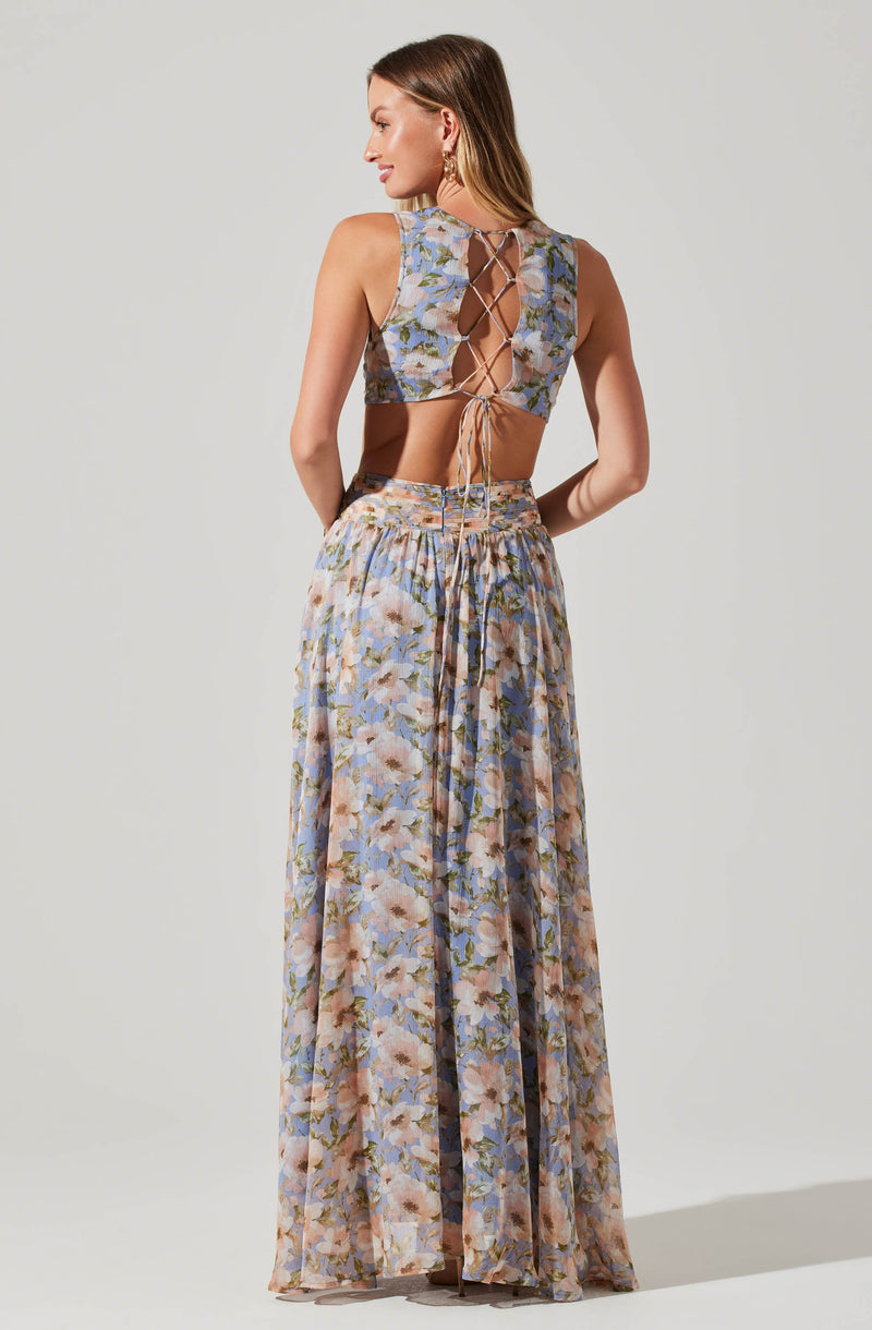 A woman is wearing a sleeveless crop top and a matching long skirt, both made of lightweight, sheer fabric with a floral pattern in shades of blue, pink, and green. The top has a lace-up back design with crisscrossing ties, while the skirt features a high waist with an elastic band and a drawstring tie at the front. The floral pattern on both pieces appears soft and delicate, creating a coordinated, elegant look.