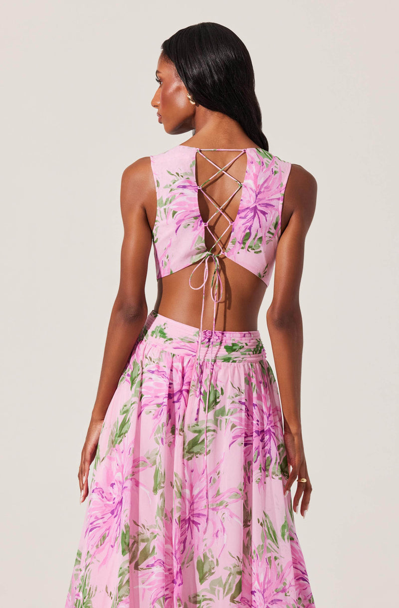 The image features a woman wearing a two-piece outfit with a light pink background and a large floral pattern in shades of pink, purple, and green. The top has a sleeveless design with a cut-out back that is laced up with thin strings crisscrossing down and tied at the bottom. The skirt is high-waisted and flowing, matching the floral pattern of the top.