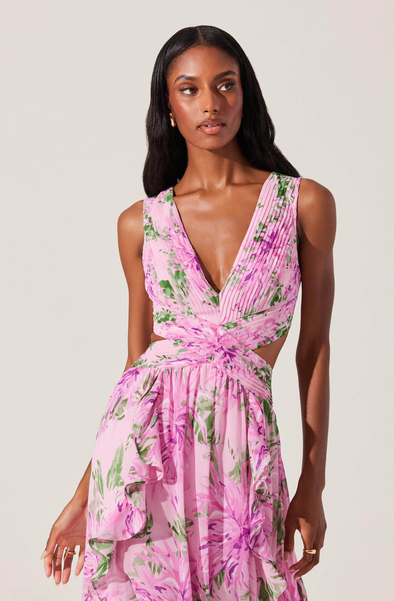 A woman is wearing a sleeveless, V-neck dress with a pink and green floral pattern. The top has a ruched design with cutouts at the waist, and the skirt is flowy with ruffled layers.