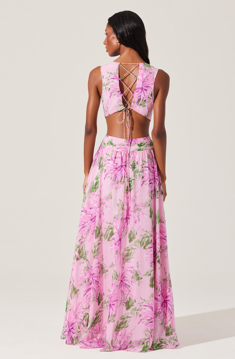 A woman is wearing a two-piece floral outfit consisting of a sleeveless crop top and a long, flowing skirt. The top features a lace-up back with crisscrossing strings, and both garments are decorated with a pink and green floral pattern on a light pink background.