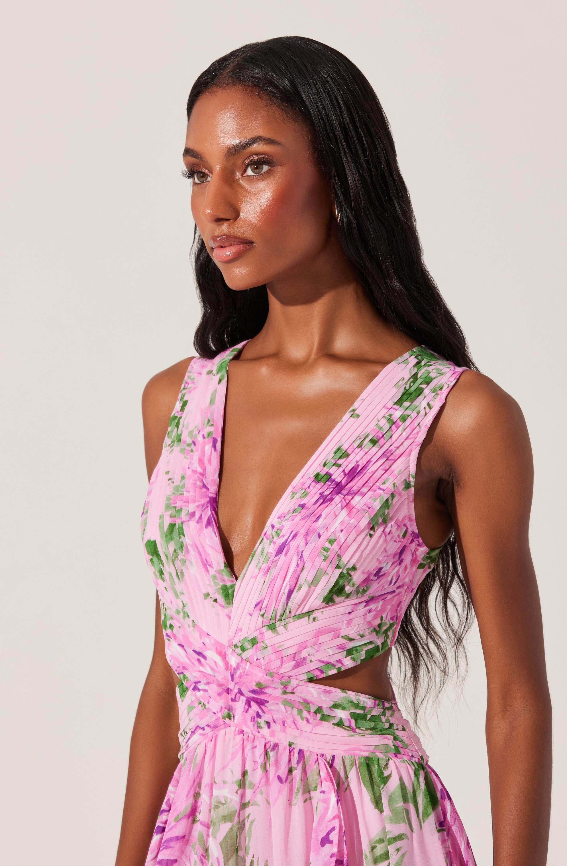 A woman is wearing a sleeveless dress with a deep V neckline. The dress features a pink base color with green and purple floral and abstract patterns. It has a fitted bodice with draped and gathered fabric, creating a cut-out at the waist, and a flowing skirt.