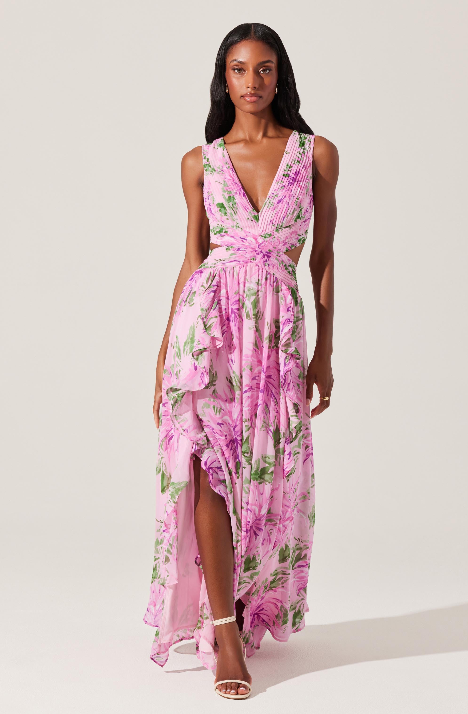 A woman is wearing a sleeveless, V-neck, floor-length dress with a pink floral print. The dress has gathered fabric at the waist, creating a fitted bodice, and features side cutouts at the waist. The skirt has a slit up the front, revealing her legs and sandals.