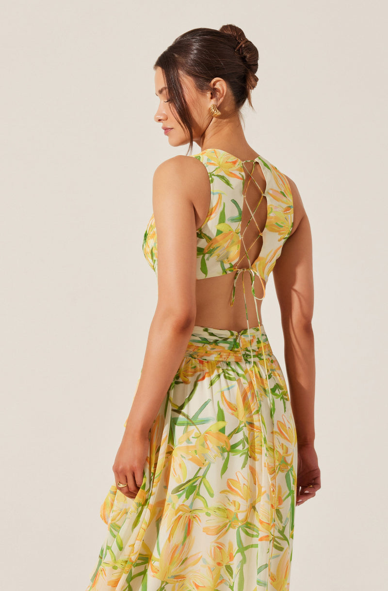 A woman is wearing a sleeveless, cropped top with a floral pattern featuring yellow and orange flowers with green leaves. The top has a lace-up back detail with thin ties. She is also wearing a matching high-waisted skirt with the same floral pattern.