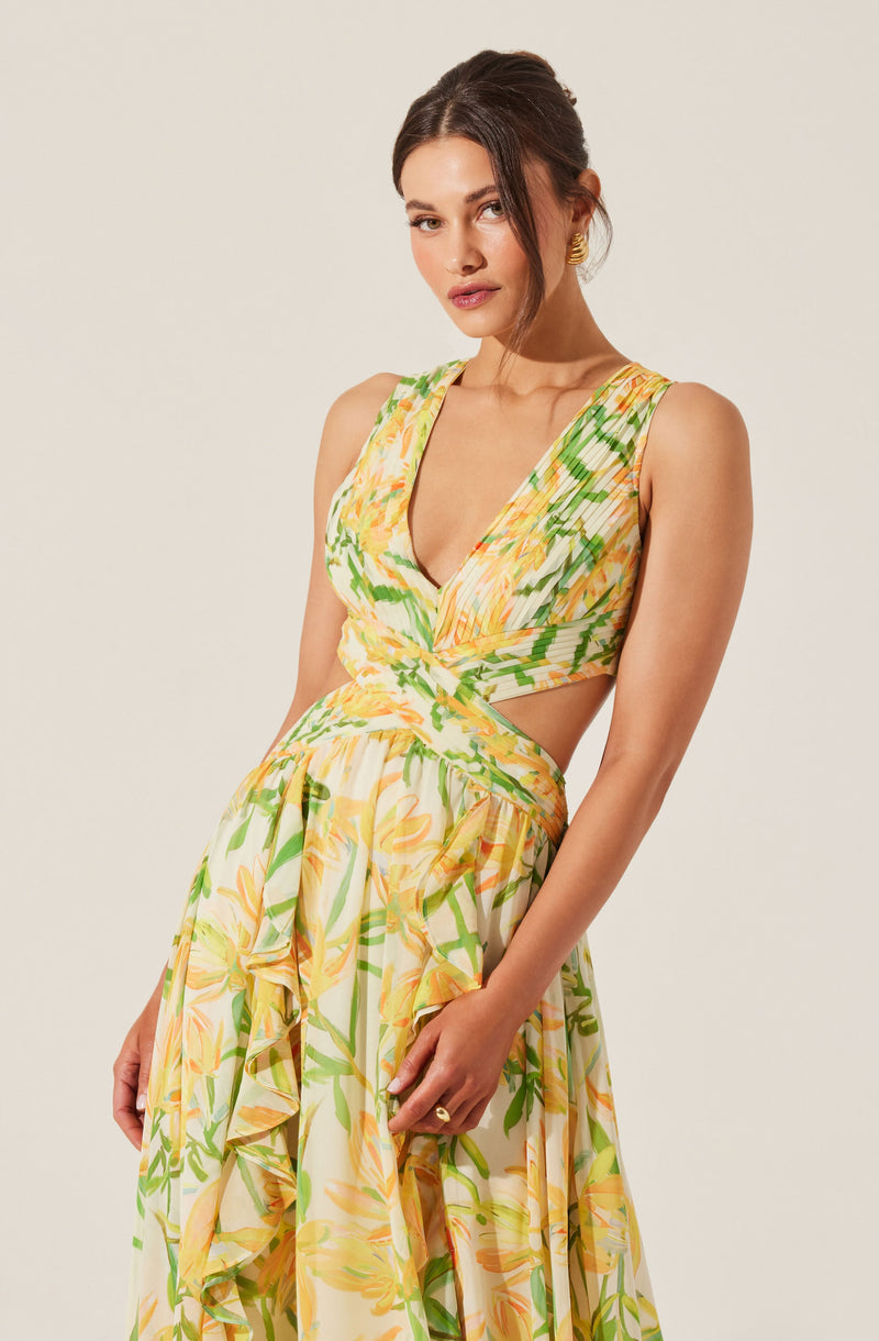 A woman is wearing a sleeveless, V-neck dress with a floral pattern featuring shades of yellow, orange, and green. The dress has cutouts at the waist, with fabric wrapping around the midsection, and a long, flowing skirt with ruffled details.