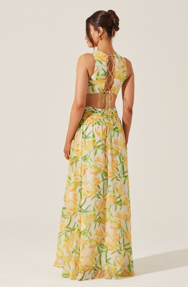 A woman is wearing a sleeveless, floor-length dress with a tropical floral pattern of yellow flowers and green leaves. The top has a cut-out back with a lace-up detail, and the fabric appears lightweight and sheer.