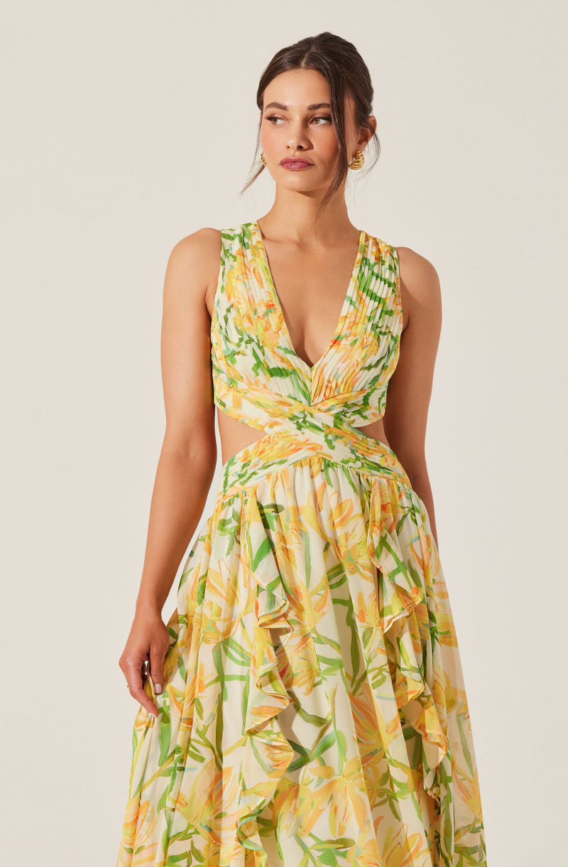 A woman is wearing a sleeveless, V-neck dress with a colorful floral pattern in shades of yellow, orange, and green. The dress features a twisted knot at the waist and ruffled layers on the skirt. The fabric appears lightweight and flowy.