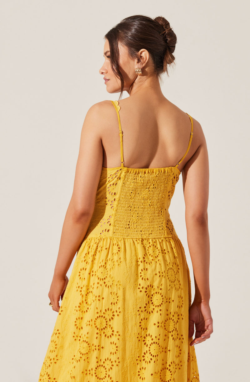 A woman is wearing a yellow dress with thin adjustable spaghetti straps. The dress features an embroidered eyelet pattern with circular and floral designs throughout, with a fitted bodice that has smocked detailing at the back. The skirt is full and gathered, creating a flowing silhouette.