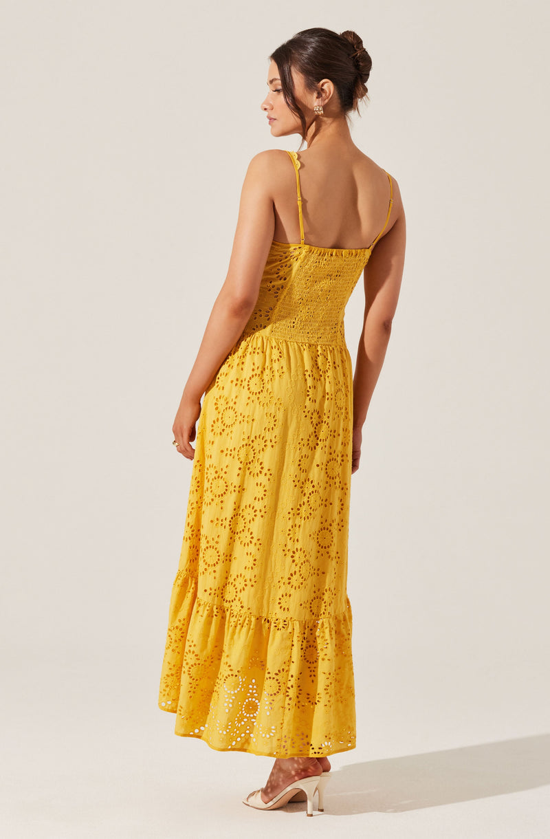 A woman is wearing a bright yellow, sleeveless summer dress with thin spaghetti straps. The dress features eyelet embroidery with circular and floral patterns throughout, and a ruffled hem at the bottom. She is wearing light-colored high heels.
