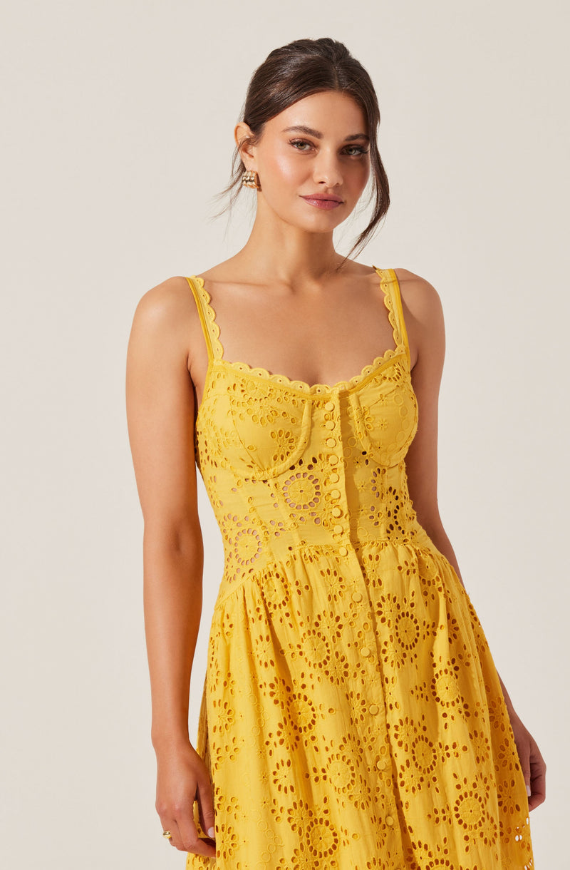 A woman is wearing a sleeveless, yellow dress made of embroidered eyelet fabric. The dress features a fitted bodice with decorative scalloped edges along the neckline and straps, and a button-down front. The skirt is gathered and flows outward, creating a soft, feminine silhouette.