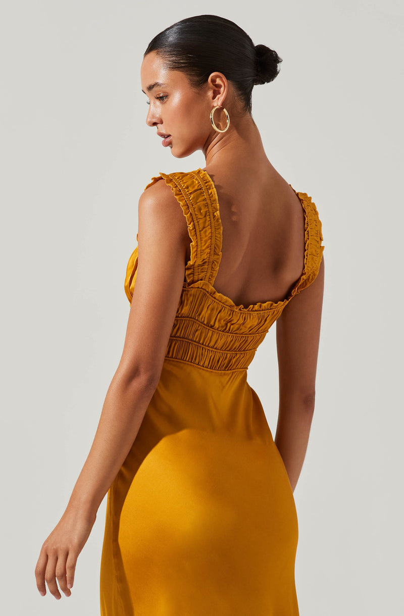 A woman wearing a sleeveless, mustard yellow dress with ruffled details on the shoulder straps and gathered fabric at the torso. The dress has a fitted waist and smooth, shiny fabric. She has her hair styled in a sleek, low bun and is accessorized with large, gold hoop earrings.