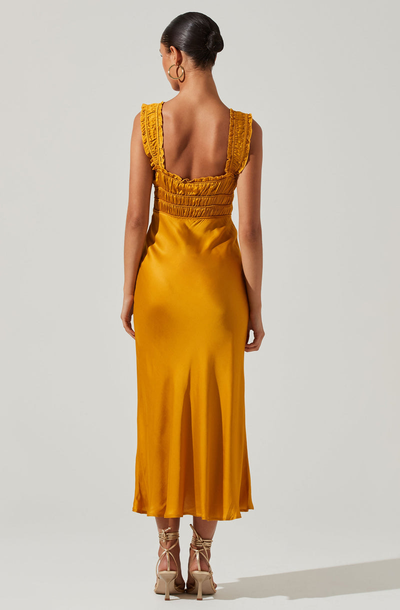 A woman is wearing a sleeveless, mustard yellow satin dress with ruffled shoulder straps and gathered detailing at the bodice. The dress is fitted at the waist and flows down to a midi length with a slight flare at the hem. She is also wearing nude, high-heeled sandals with ankle straps.