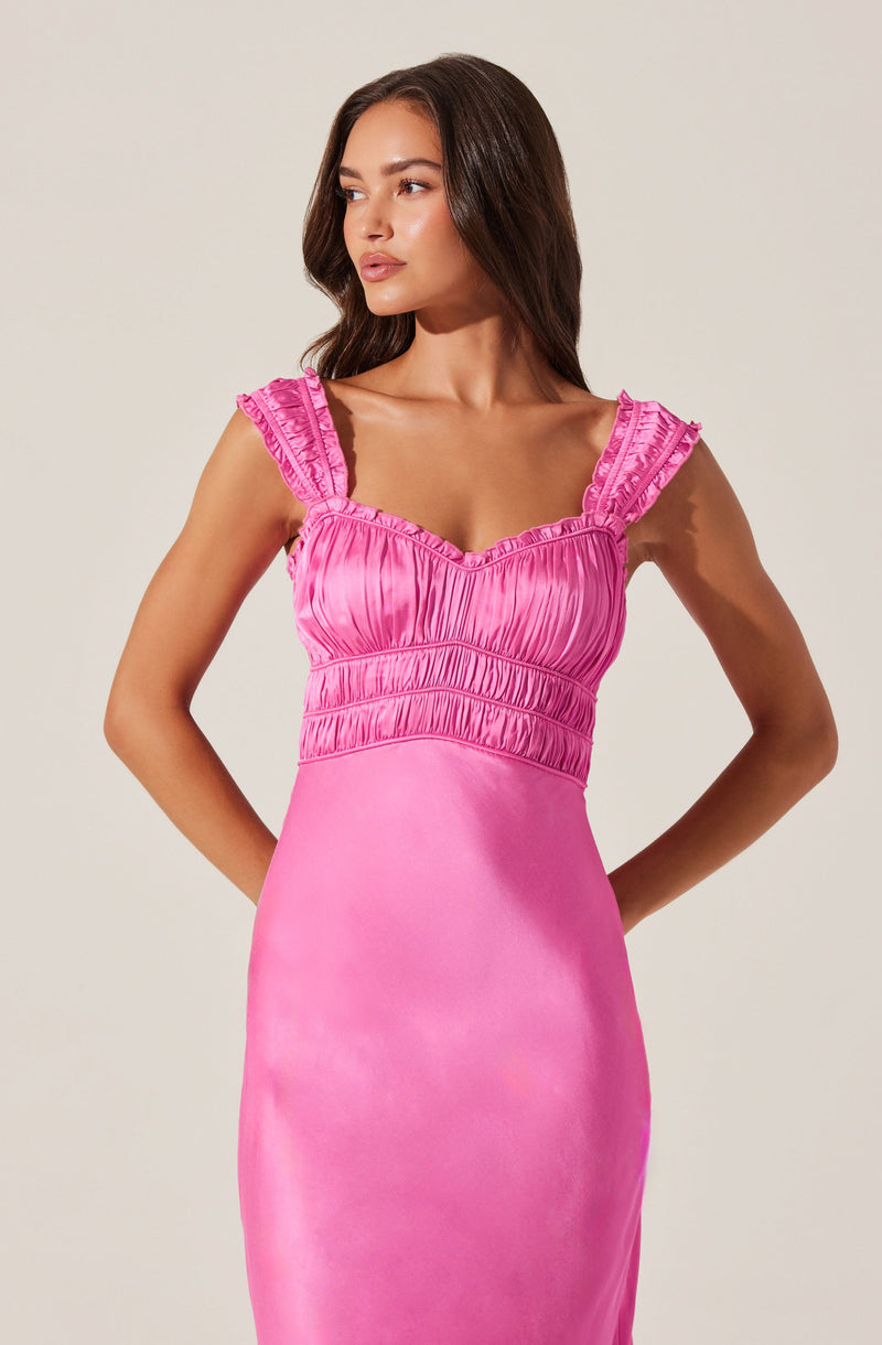 A woman is wearing a sleeveless, pink dress with ruffled shoulder straps, a ruched bodice with vertical pleats, and a smooth, fitted skirt.