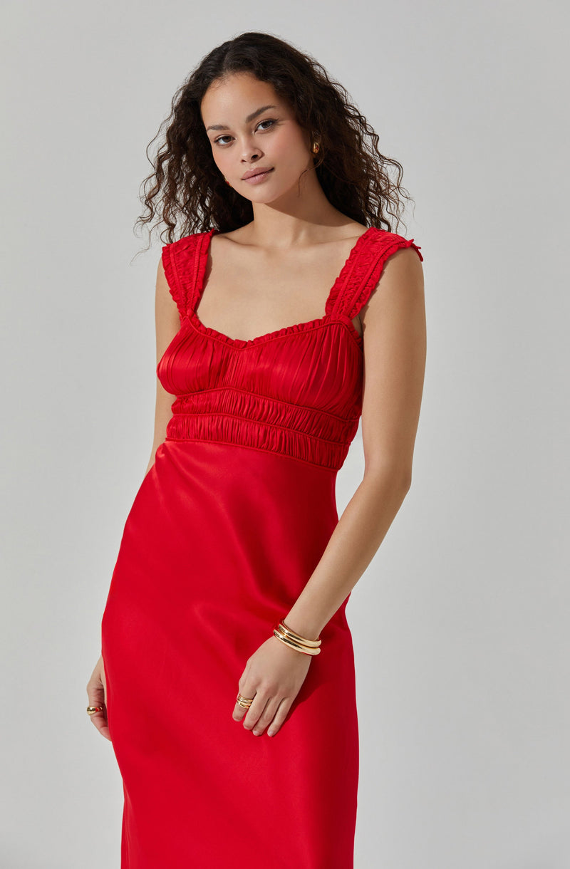 The woman is wearing a bright red sleeveless dress with gathered and textured fabric details around the bodice and shoulder straps. The dress has a fitted waist and a smooth, flowing skirt. She has gold jewelry, including earrings, bracelets, and rings.