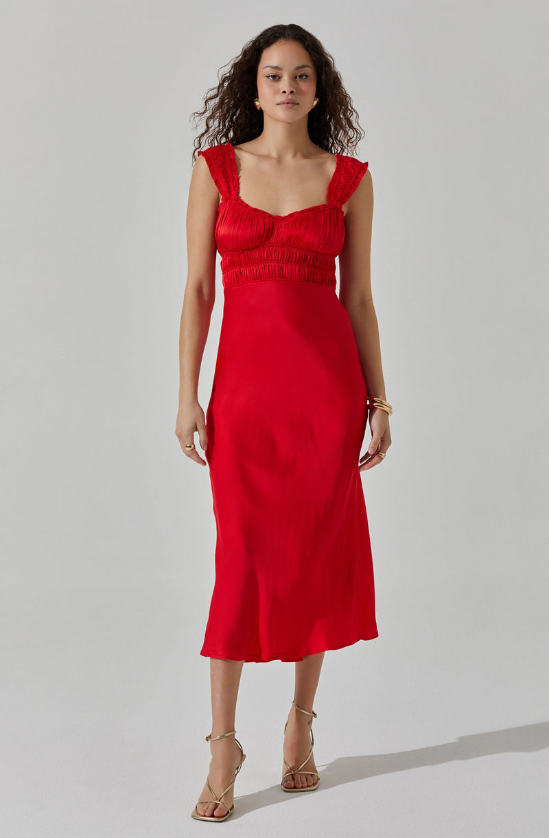 A woman is wearing a sleeveless, red, midi-length dress with gathered details on the bodice and ruffled straps. She is accessorized with gold earrings, a bracelet, and rings, and is wearing nude, strappy high-heeled sandals.