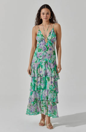 A woman is wearing a sleeveless, long summer dress with spaghetti straps. The dress features a vibrant floral pattern in shades of green, purple, and lavender, with a keyhole cutout at the front. It has a tiered, ruffled skirt that reaches down to her ankles.