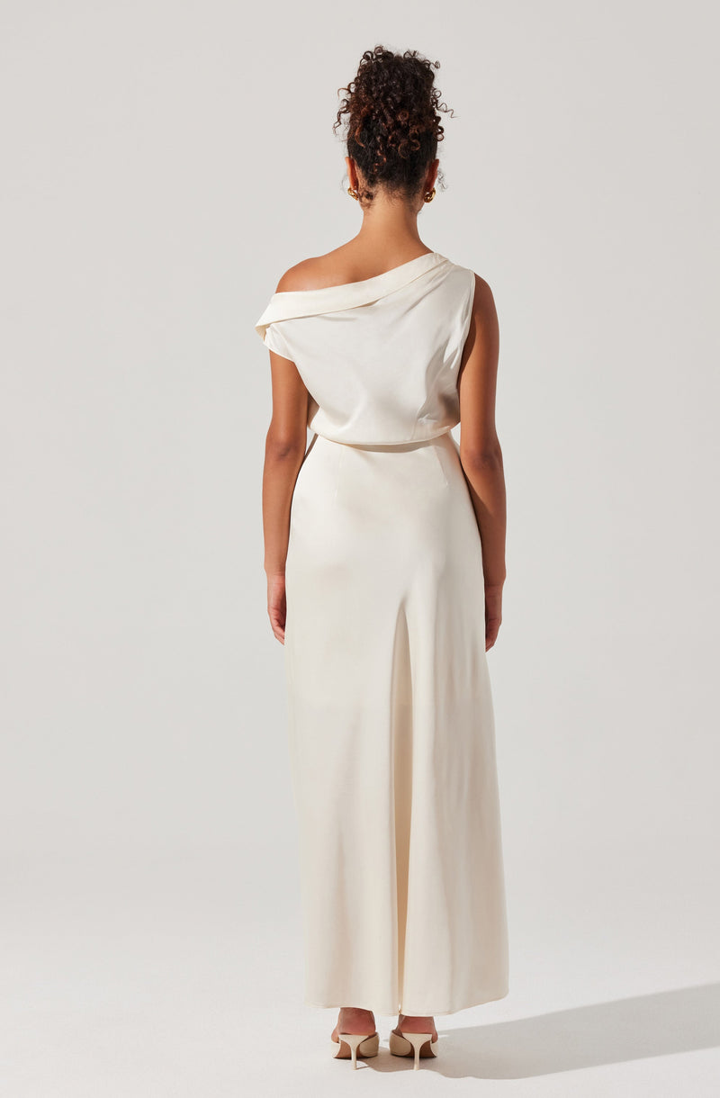 The woman is wearing a sleeveless, off-shoulder, white dress with a draped neckline and a high waist. The dress features a long, flowing skirt with a slit at the back. She is also wearing light-colored high-heeled shoes.