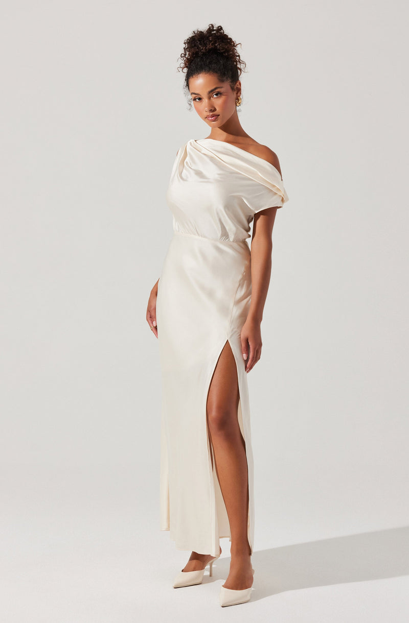 A woman is wearing an elegant off-the-shoulder floor-length gown in a shiny satin fabric. The dress features a draped bodice that falls loosely over her shoulders and a high slit on the side, revealing her leg. She completes the look with a pair of pointed-toe heels in a matching light color.