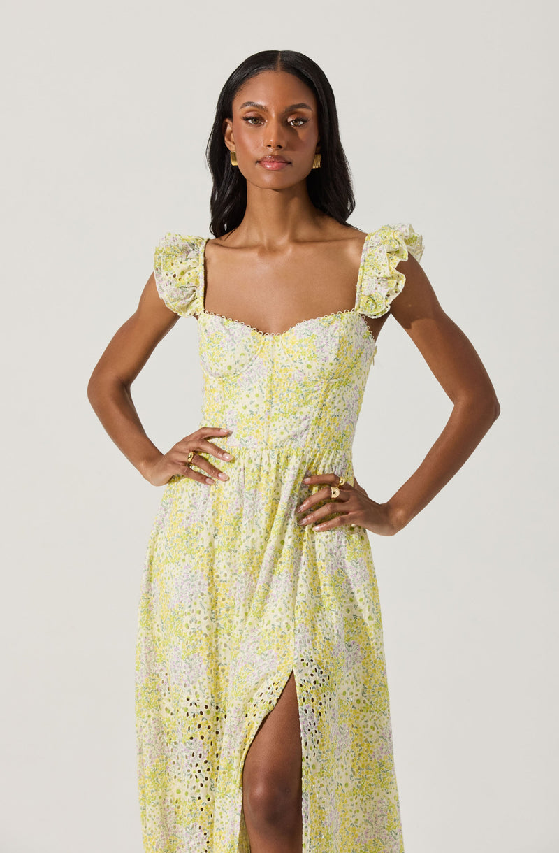 A woman is wearing a light yellow and white floral dress with small multicolored flowers. The dress has ruffled, capped shoulder sleeves and a fitted bodice with a sweetheart neckline. The skirt is long, flowing, and features a slit up the front, revealing her leg.