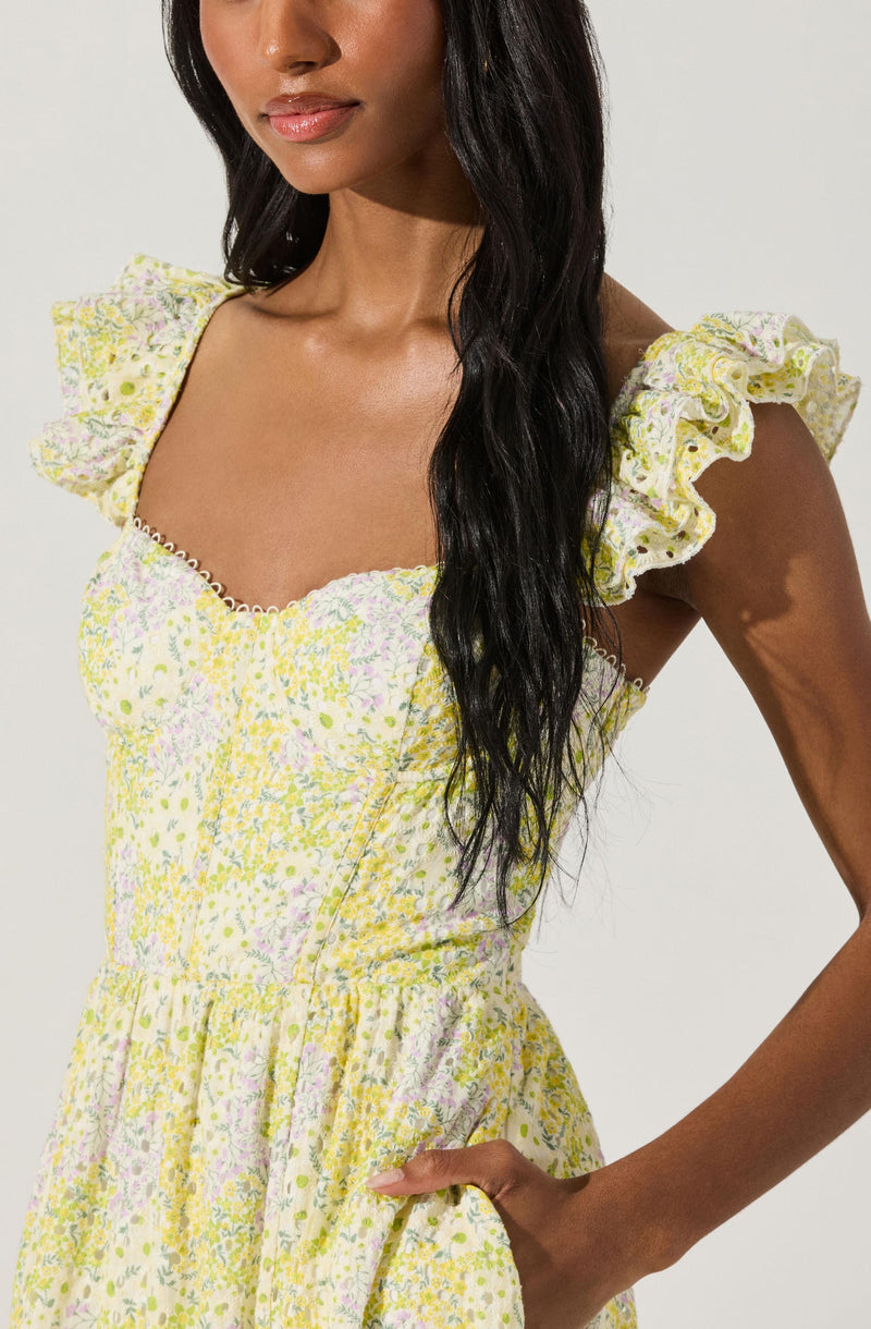 A woman is wearing a light-colored dress with a floral pattern in shades of yellow, pink, and green. The dress has a fitted bodice with a sweetheart neckline bordered by a delicate trim, and ruffled shoulder straps. The fabric appears lightweight and airy, suitable for warm weather.