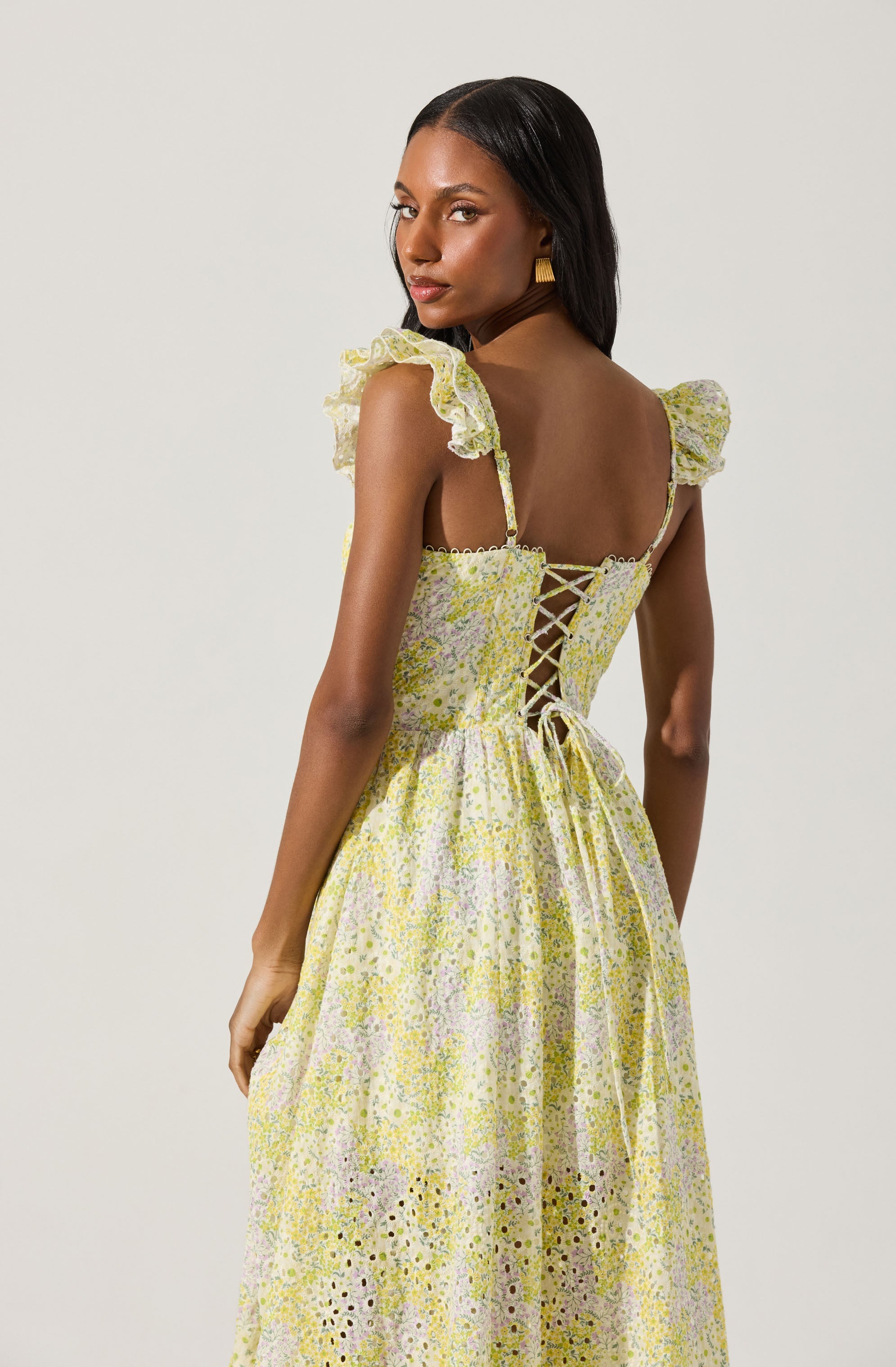 A woman is wearing a long, light yellow floral dress with ruffled shoulder straps and a lace-up back. The dress features a delicate pattern of small flowers in shades of yellow, purple, and green, with eyelet details near the hem.
