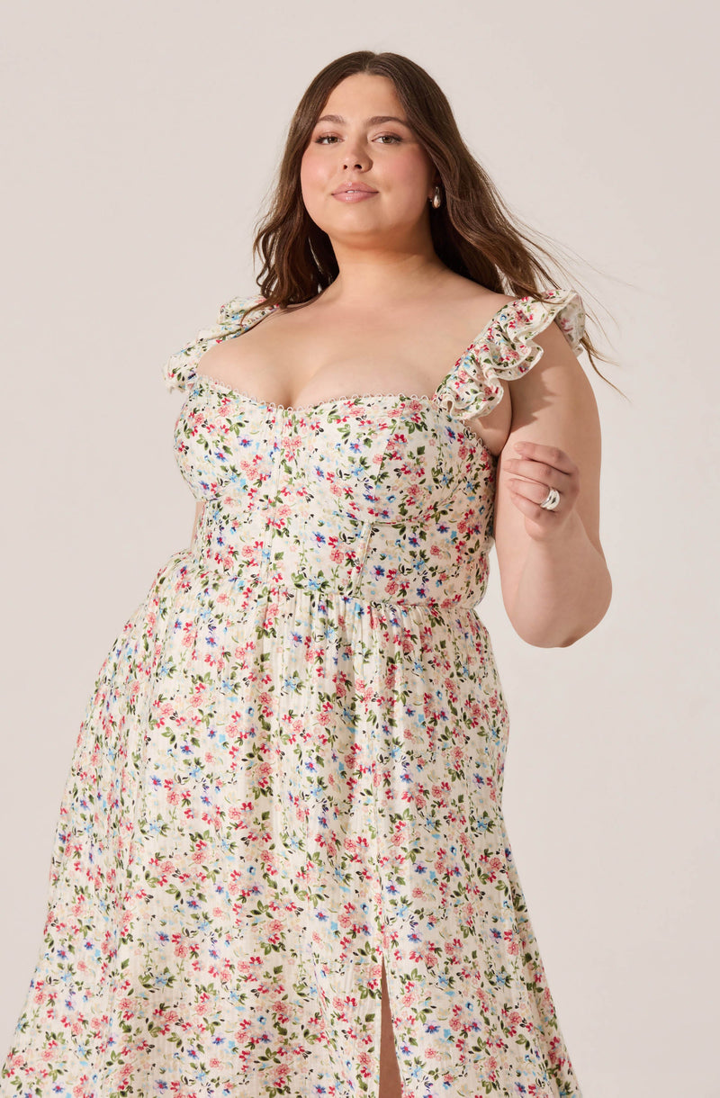 A woman wearing a sleeveless, floral print dress with ruffled shoulder straps. The dress is fitted at the waist and features a slit in the skirt. The floral pattern includes small pink, blue, and green flowers on a light background.