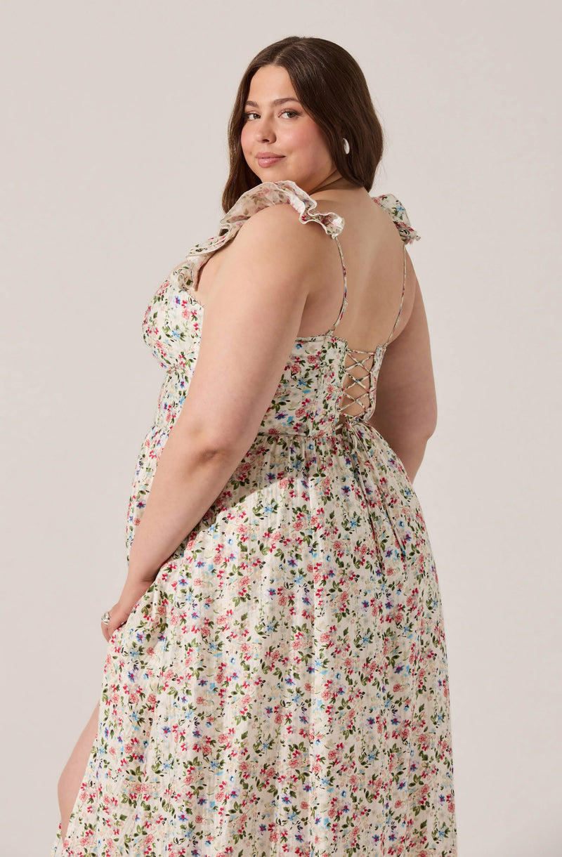 A woman is wearing a sleeveless, floral-patterned dress with ruffled shoulder straps. The dress features a lace-up detail on the back with thin straps. The fabric has a cream background with small, colorful flowers scattered throughout.