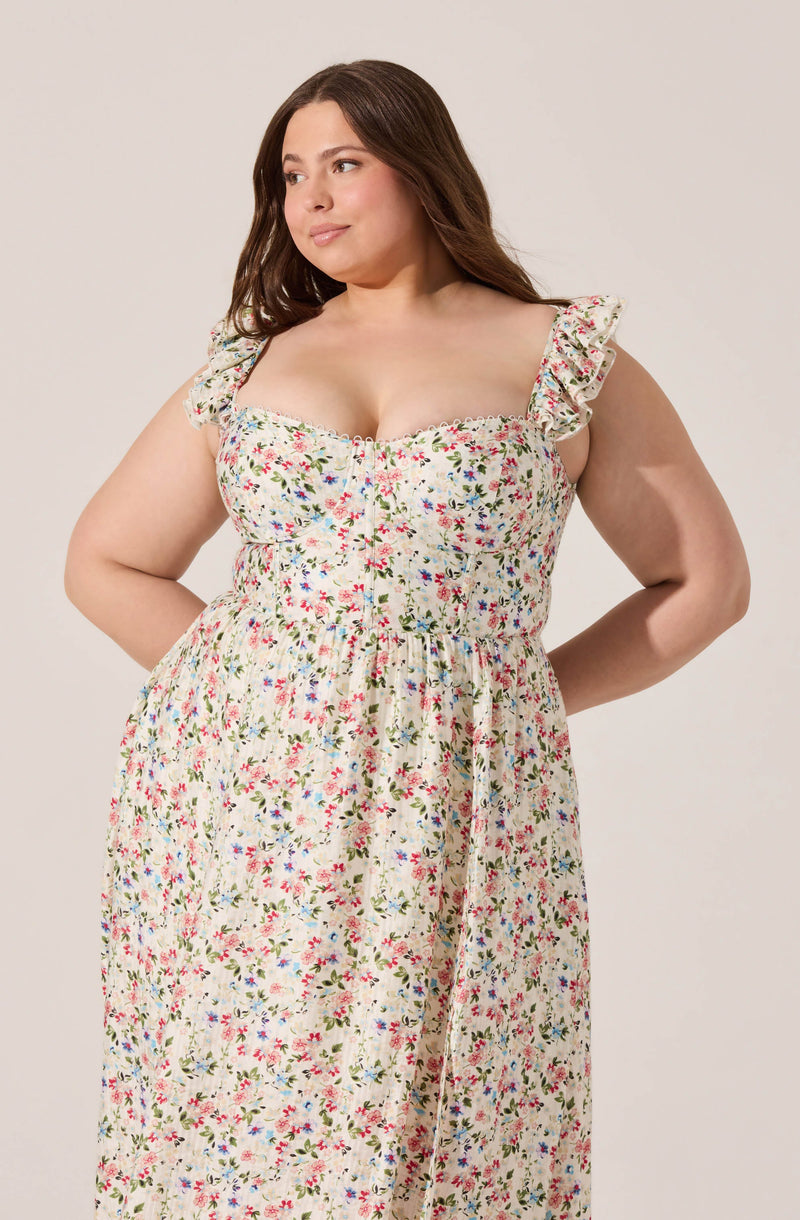 A woman wearing a long, sleeveless dress with ruffled straps and a floral pattern featuring small pink, red, blue, and green flowers on a light background. The dress has a fitted bodice with a slight V neckline and a gathered waist, flowing down to the floor.